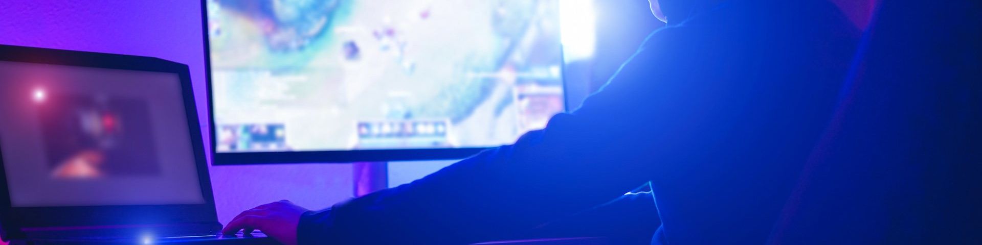 A blue/purple image of a gamer, photographed from the side and behind. They sit in a high backed chair, facing two computer screens.